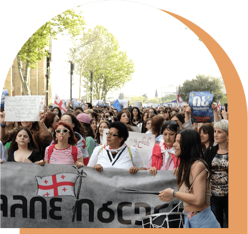 A large group of people stand together outdoors at a protest. In the foreground, several people are holding a large gray banner that includes a Georgian flag. Many people in the rest of the crowd are also carrying signs and/or Georgian flags. All of the text on the signs is in Georgian.