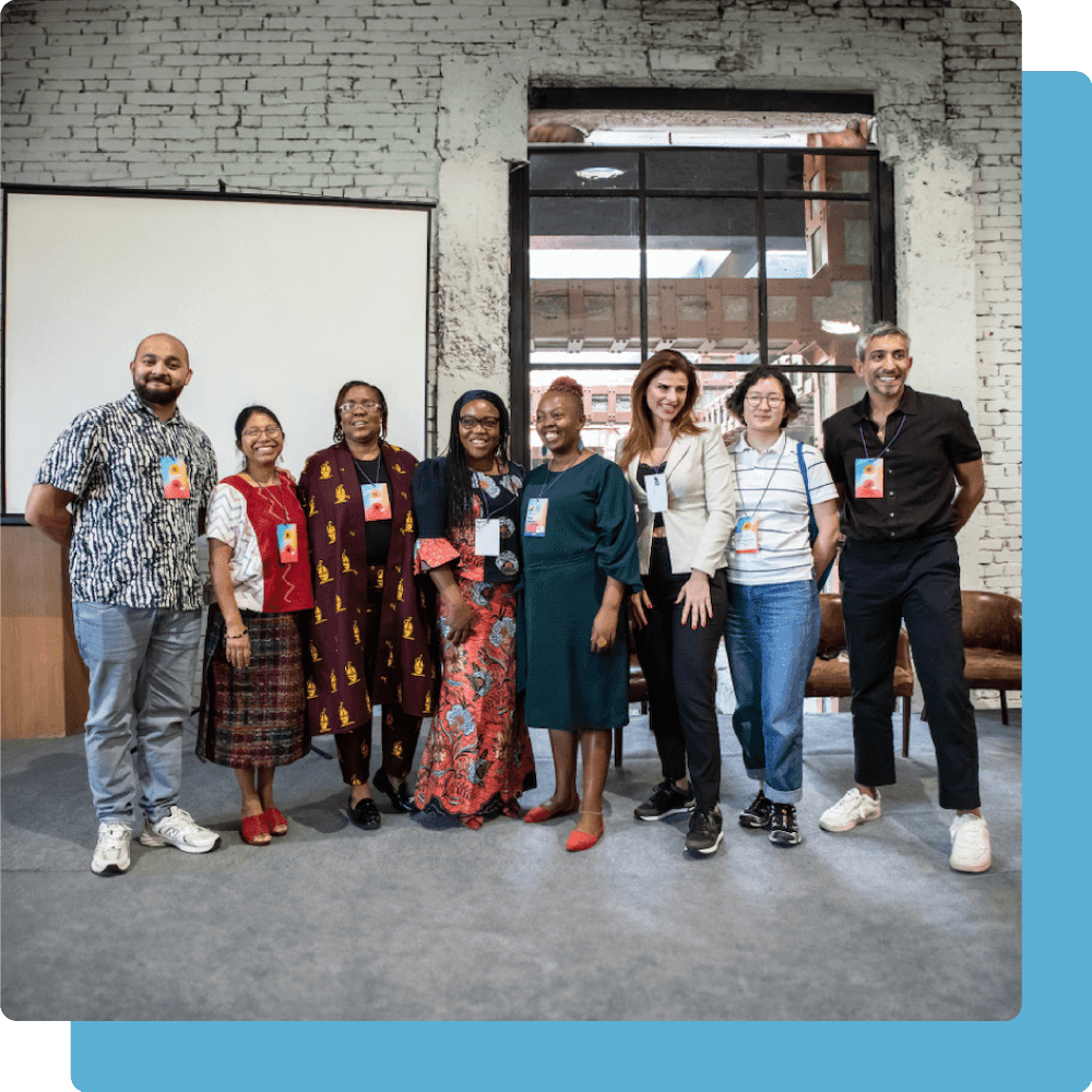 A group of eight people of varying races and gender presentations stand together in a line facing the camera. Many of them are smiling. Behind them is a projection screen.