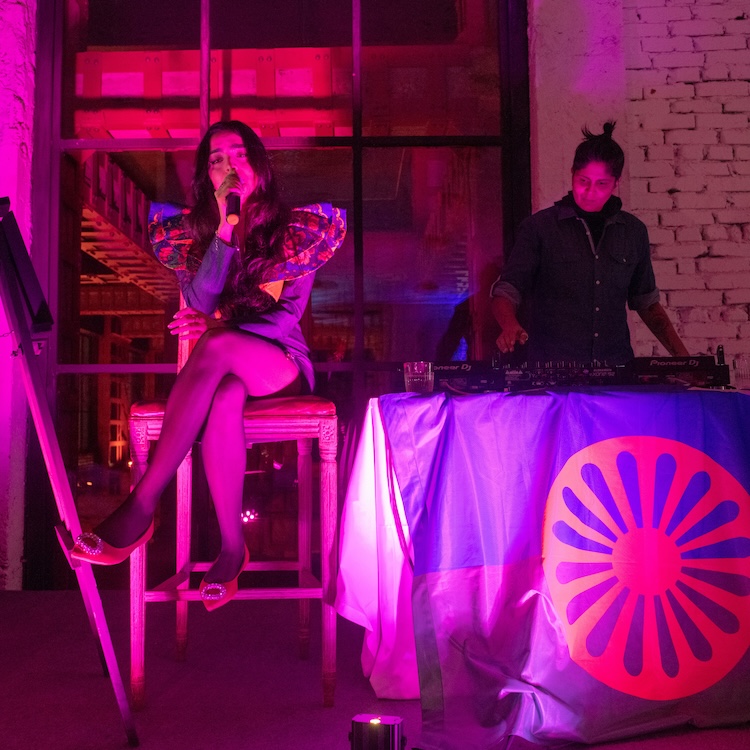 A person in a dress with puffy shoulders sits on a tall seat. Their eyes are closed and they are holding a microphone, in the middle of speaking or singing. Behind them, a second person in a button up denim shirt stands at a DJ table. The lighting is pink, casting a glow on the contents of the photo.