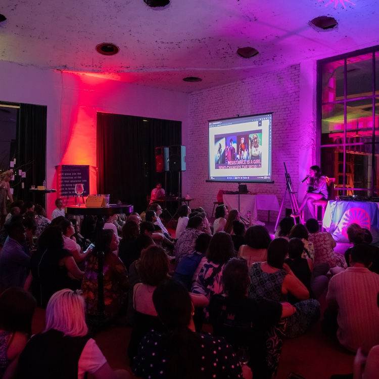 A large group of people are seated together facing a stage, where someone is seated with a microphone. There is colorful lighting, casting pink, purple, and orange glows over parts of the photo.