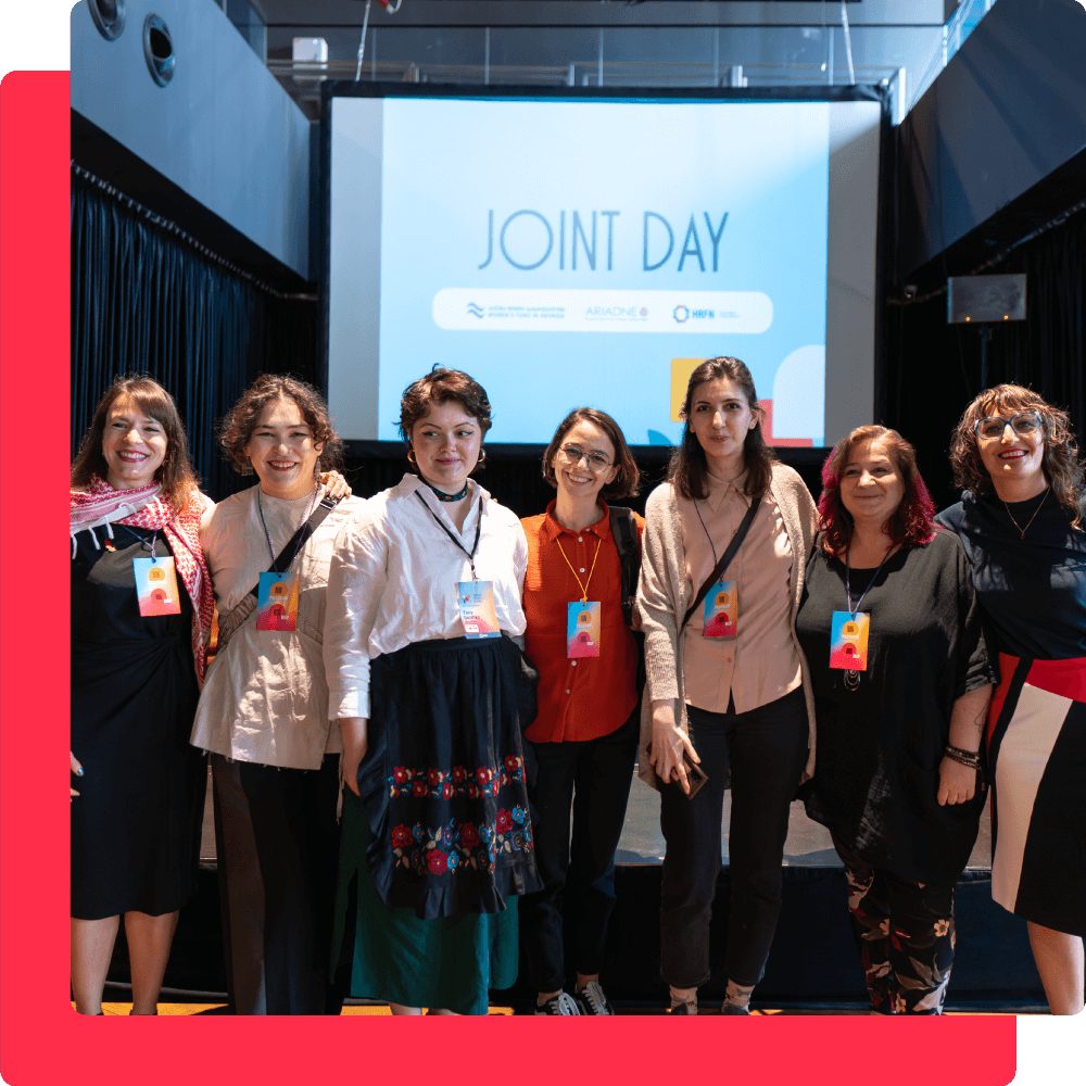 Several people stand side-by-side facing the camera, their arms around each other. Many of them are smiling. Behind them is a presentation screen that says “Joint Day” in large letters.