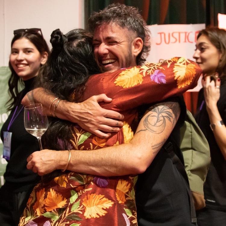 In the foreground, two people are hugging. The one facing the camera has a big smile. In the background, an onlooker is facing the huggers and smiling. A sign taped to the wall in the background says the word “Justice.”