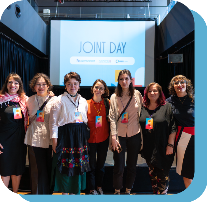Several people stand side-by-side facing the camera, their arms around each other. Many of them are smiling. Behind them is a presentation screen that says “Joint Day” in large letters.