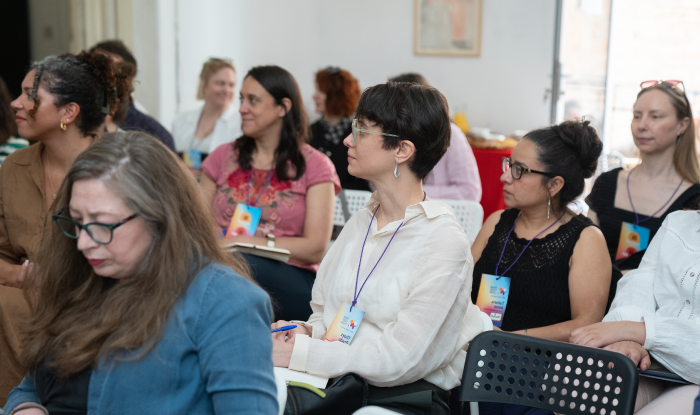 People of varying races and gender presentations are seated in rows in a conference room. They are all looking off to the side and appear to be listening closely.