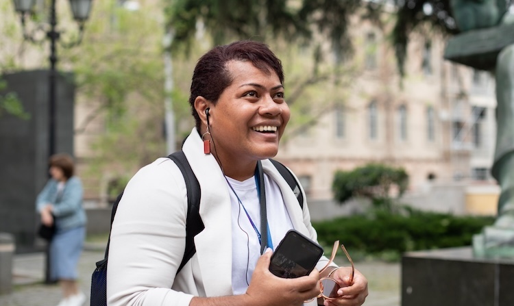 A person in a white shirt and blazer stands outside. They are holding sunglasses and a phone, and have headphones in their ears. They are looking off to the right and smiling, their expression full of emotion.