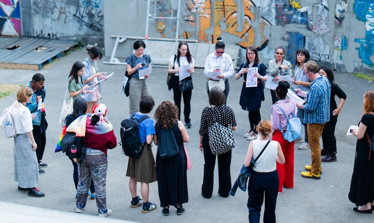 A group of people of varying races and gender presentations stands outside on concrete, colorful graffiti covering the walls around them. They are standing roughly in a circle, and appear to be engaged in a conversation.
