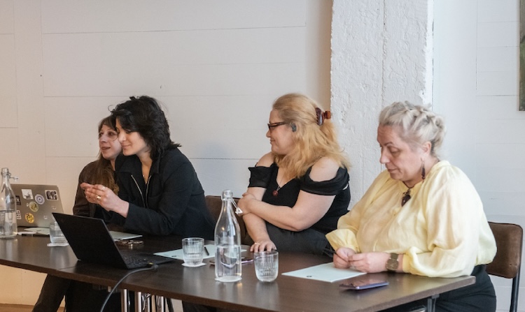 Four people sit together at a table with laptops and glasses of water in front of them. One of them appears to be facing an audience and speaking, while the other three are looking at the person speaking or down at the table, listening.