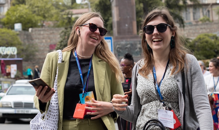 A close up of two people wearing sunglasses and conference lanyards. They are outside and appear to be walking. Both have headphones in their ears, and they are smiling.